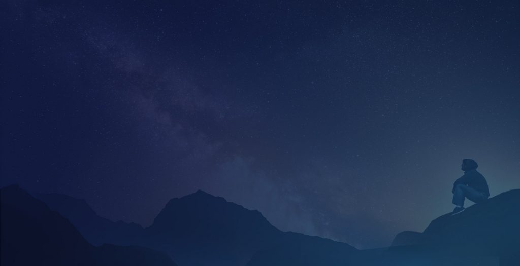 A man is sitting on top of a mountain looking at the night sky.